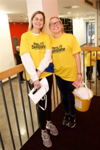 Two white females smiling at the camera, both wearing yellow Rays of Sunshine t-shirts and holding fundraising buckets