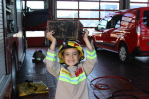 Small boy dressed in a firefighter's uniform and hat, holding up a piece of firefighting equipment and smiling for the camera. His uniform reads Khaliq.
