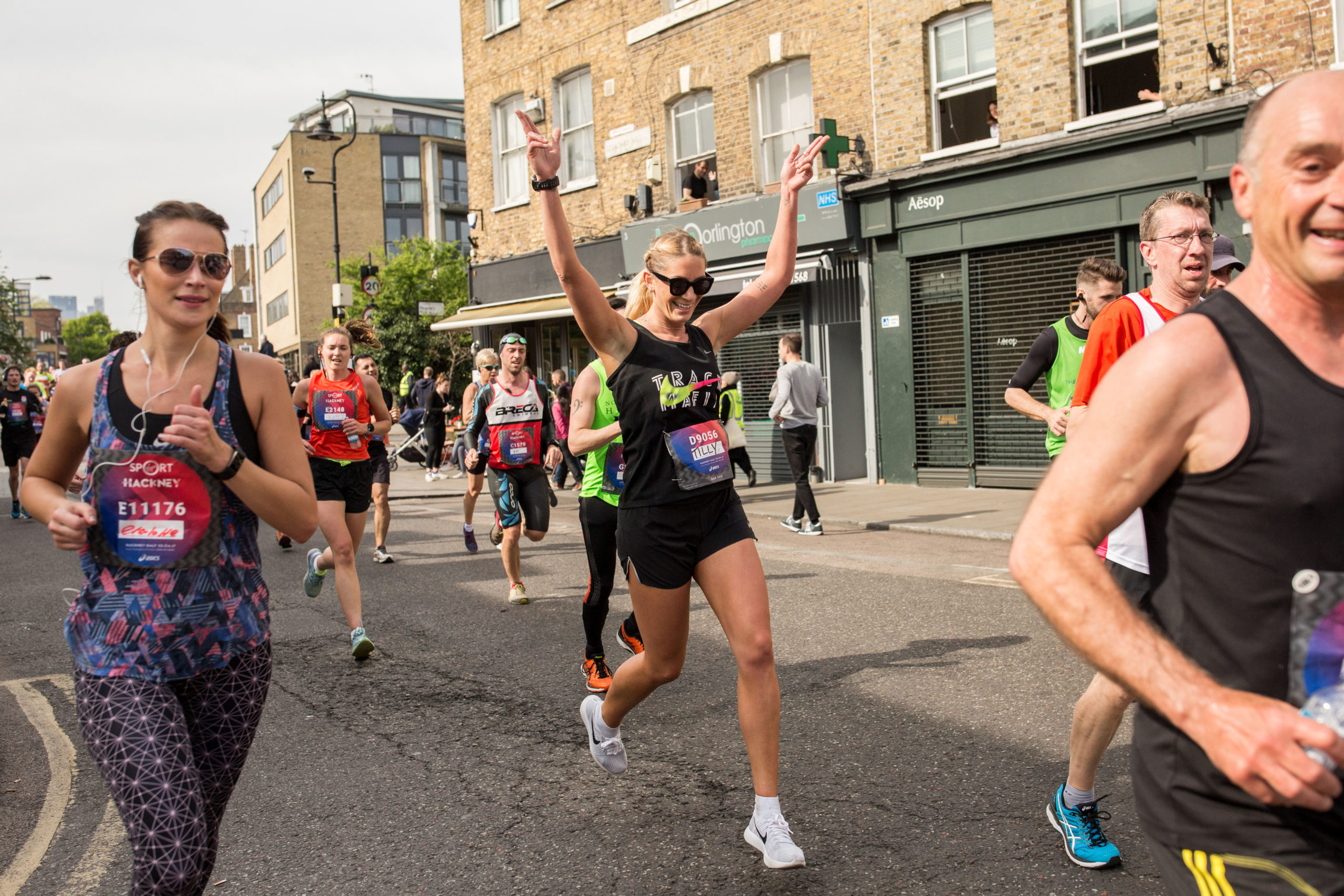 A lady in a black vest running in a street, holding her hands up while surrounded by other runners.