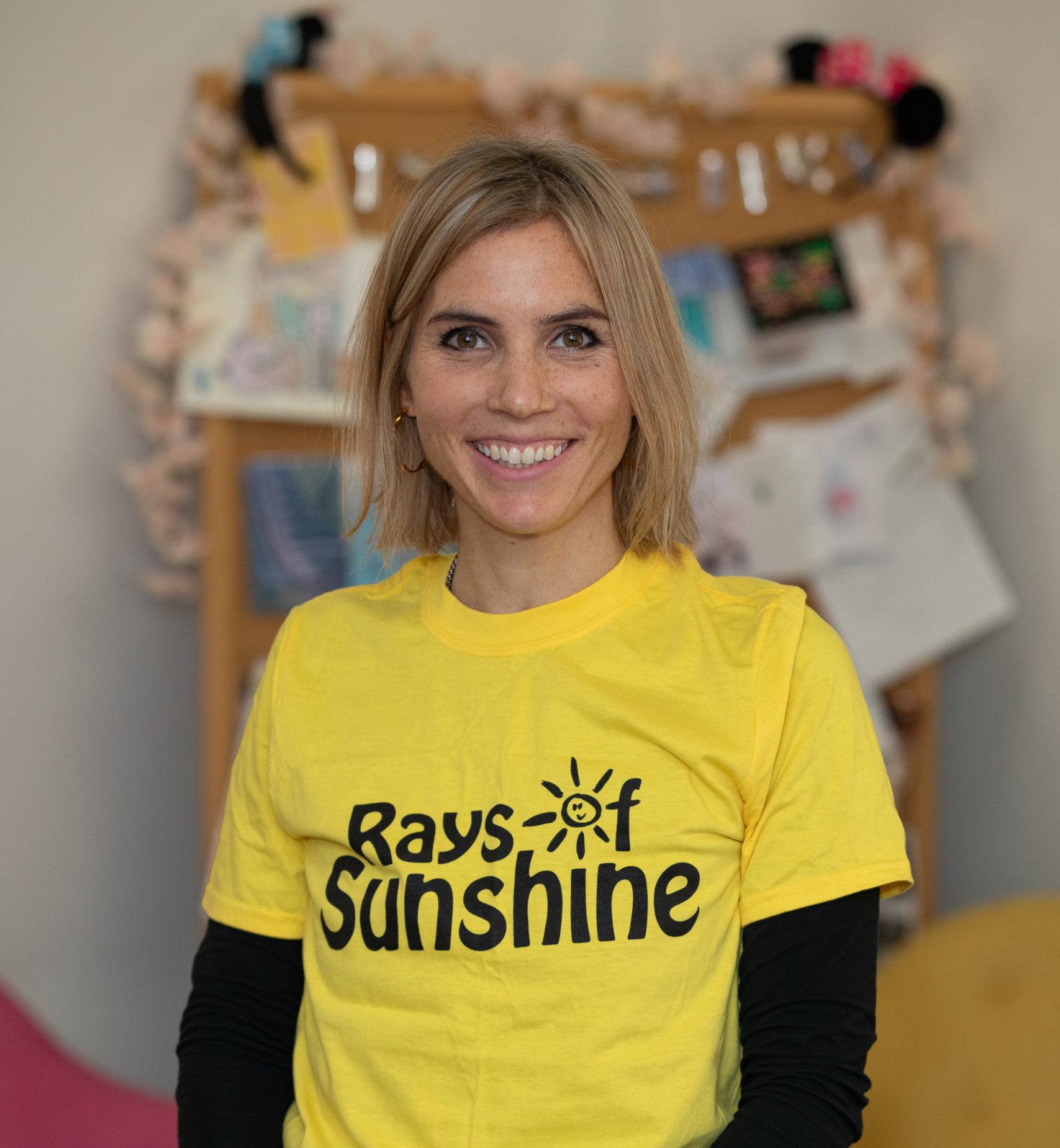 Jenny is standing and is wearing a Rays of Sunshine t-shirt. She has blonde hair.
