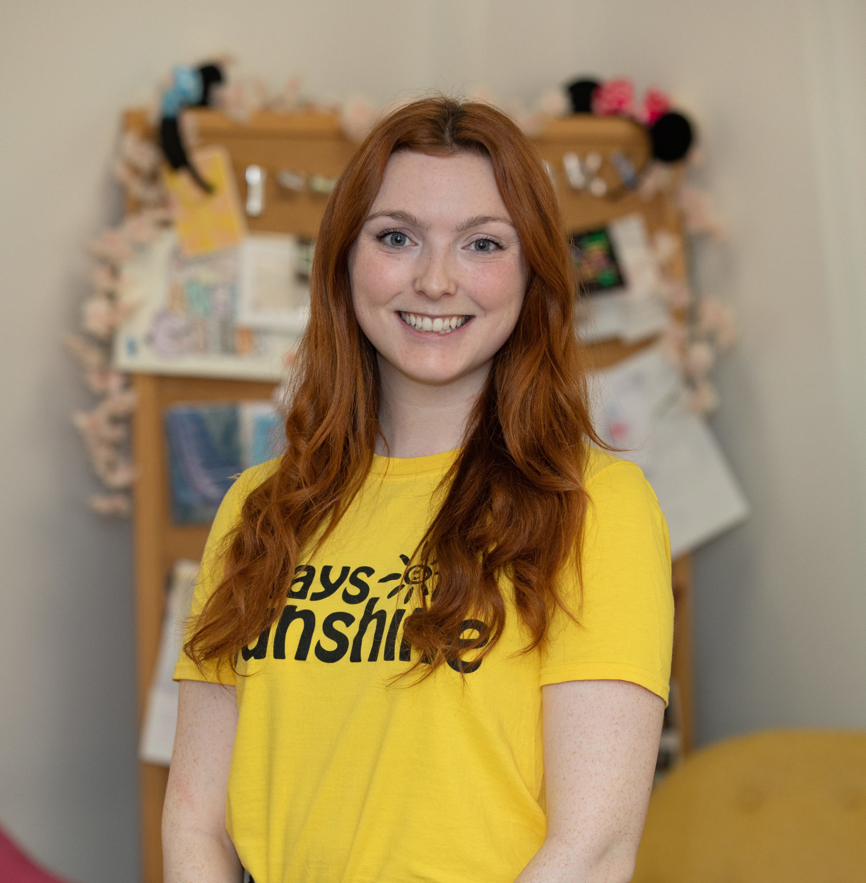 Clodagh is wearing a yellow Rays of Sunshine t-shirt. She is standing. Clodagh has ginger hair