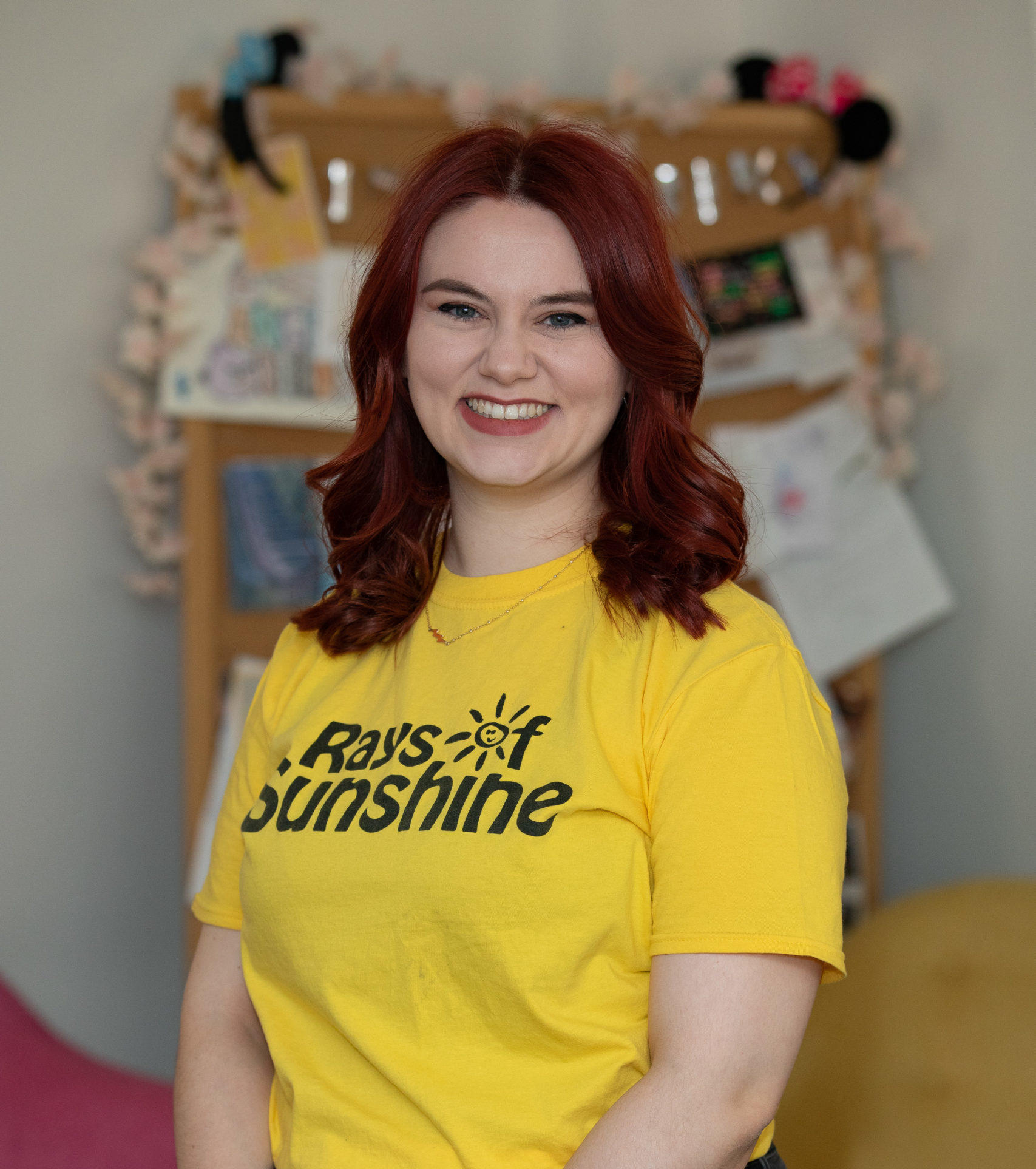 Kathryn is wearing a yellow Rays of Sunshine t-shirt. Kathryn is standing and has red hair,