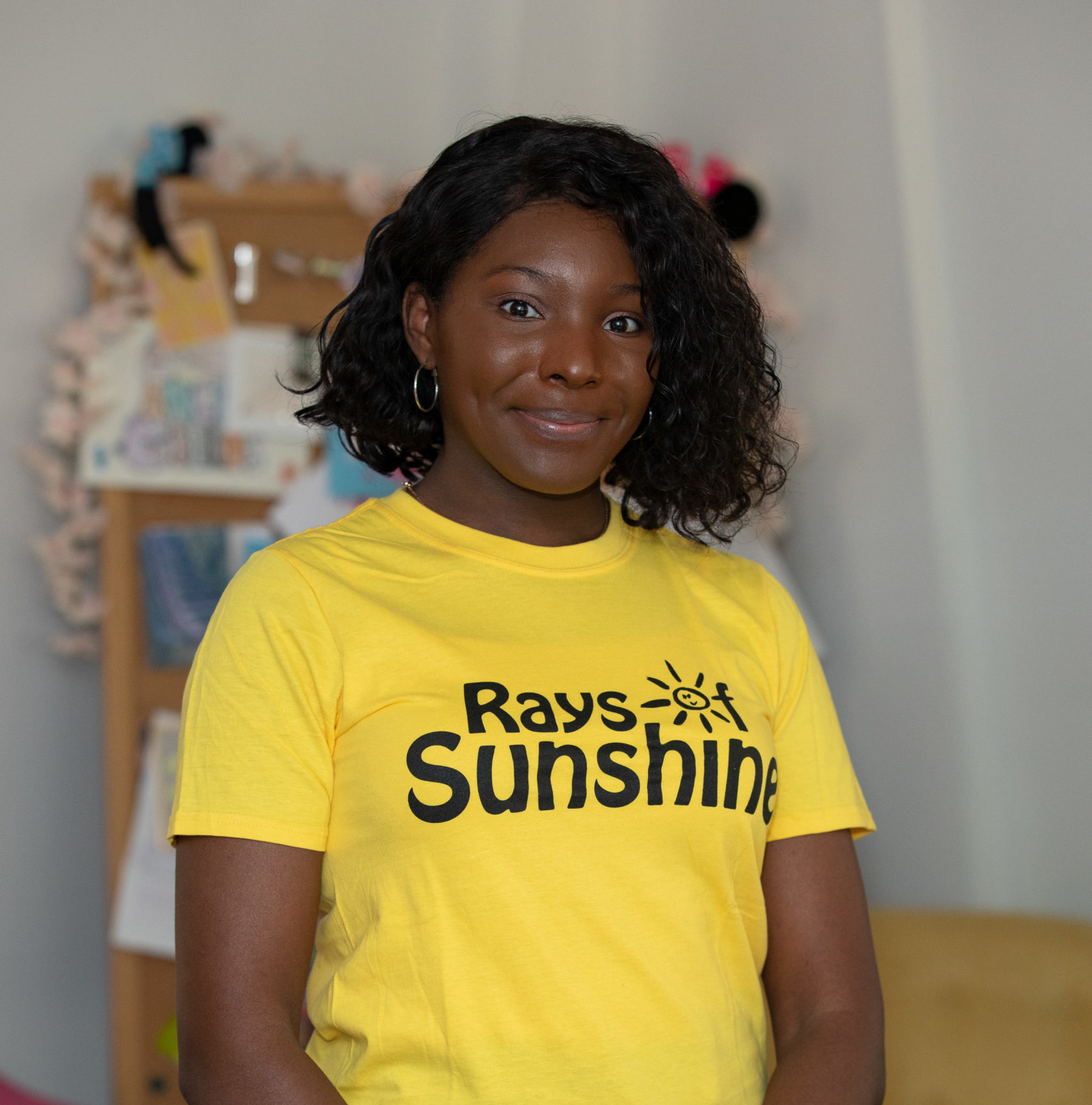 Jess is wearing a yellow Rays of Sunshine t-shirt. She is standing and has dark hair.