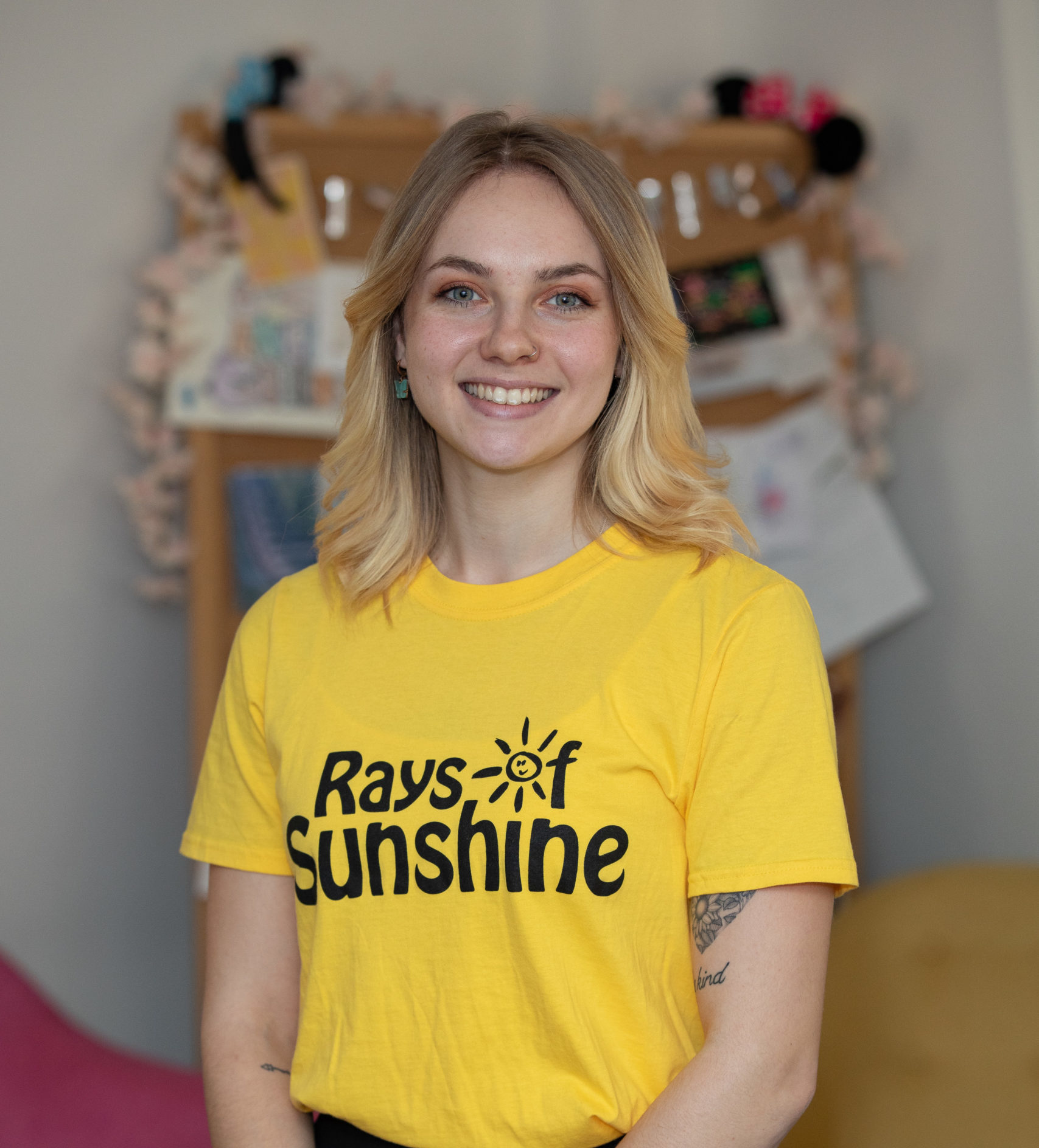 Jess is wearing a yellow Rays of Sunshine t-shirt. Jess has short blonde hair.