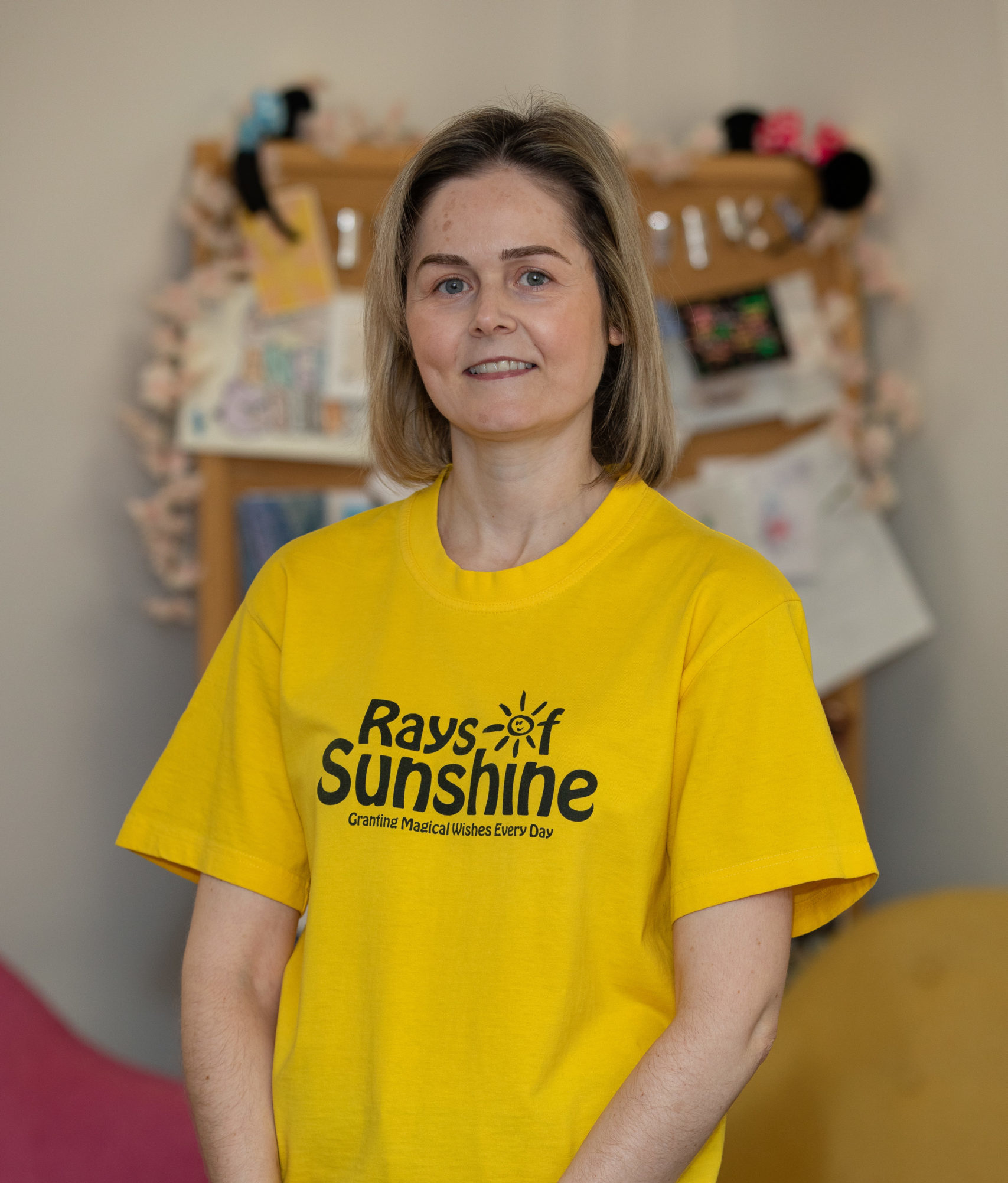 Sarah is wearing a yellow Rays of Sunshine t-shirt. She is standing.