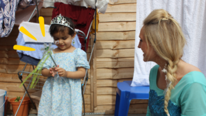 A little girl with a feeding tube is playing with a magic wand. She is next to a blonde woman dressed as Elsa.