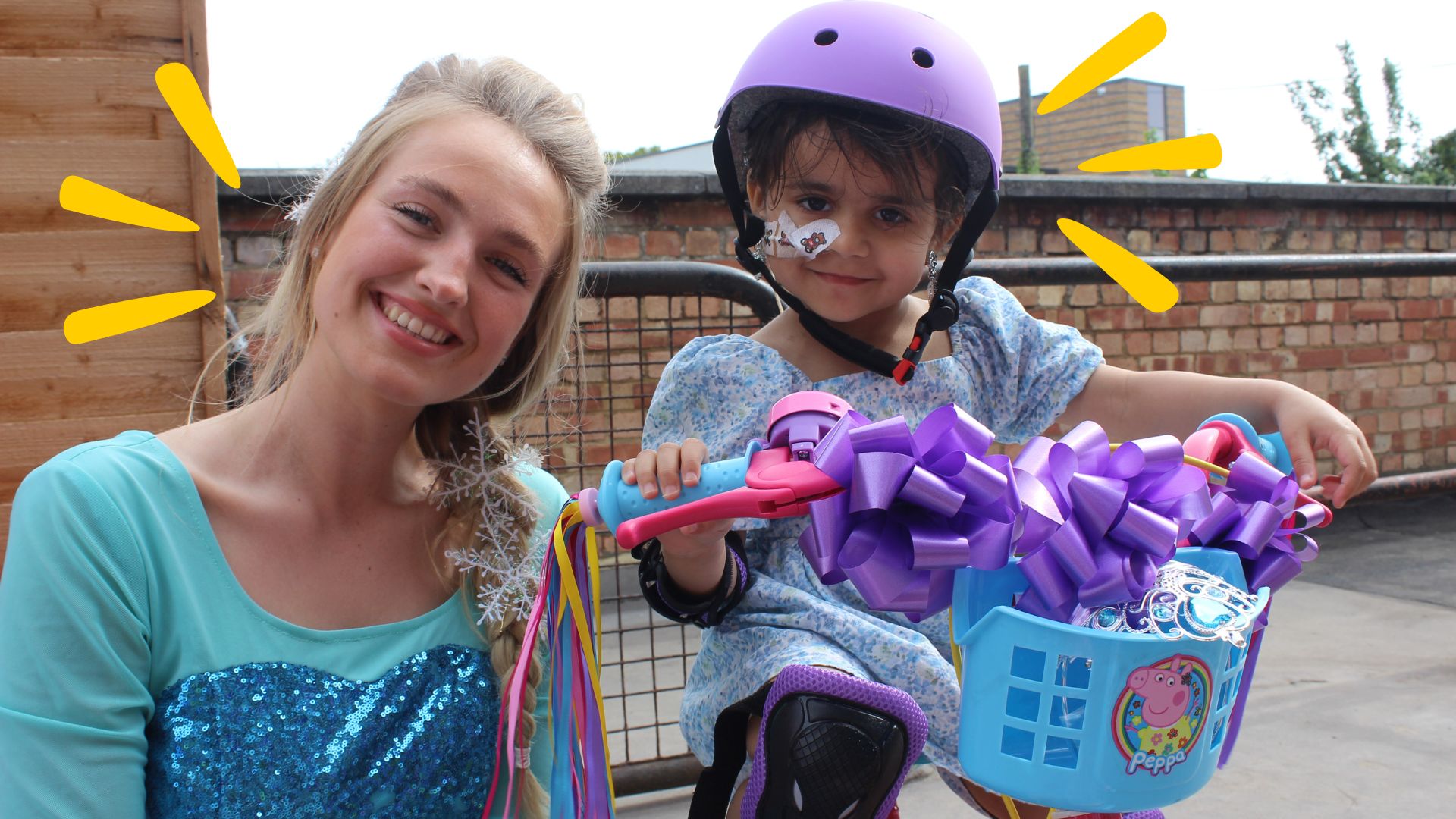 A little girl with a feeding tube is riding a Peppa Pig bike which is adorned with a big purple bow. She is next to a blonde woman dressed as Elsa.