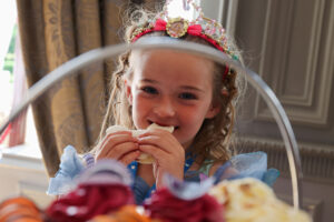 Little girl wearing a tiara smiling while eating a sandwich