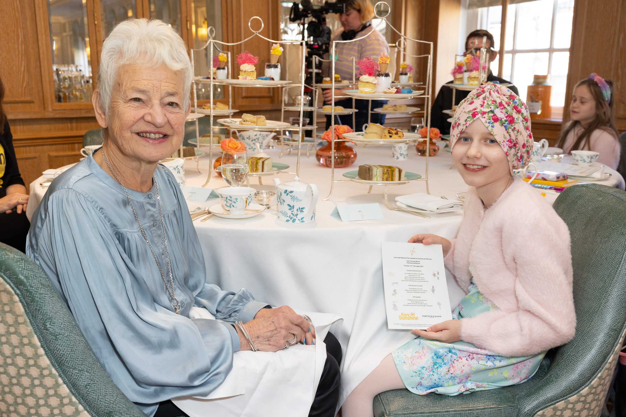 Little girl in pink cardigan and floral headscarf sits next to and smiles with older woman with short white hair and a silky blue shirt.