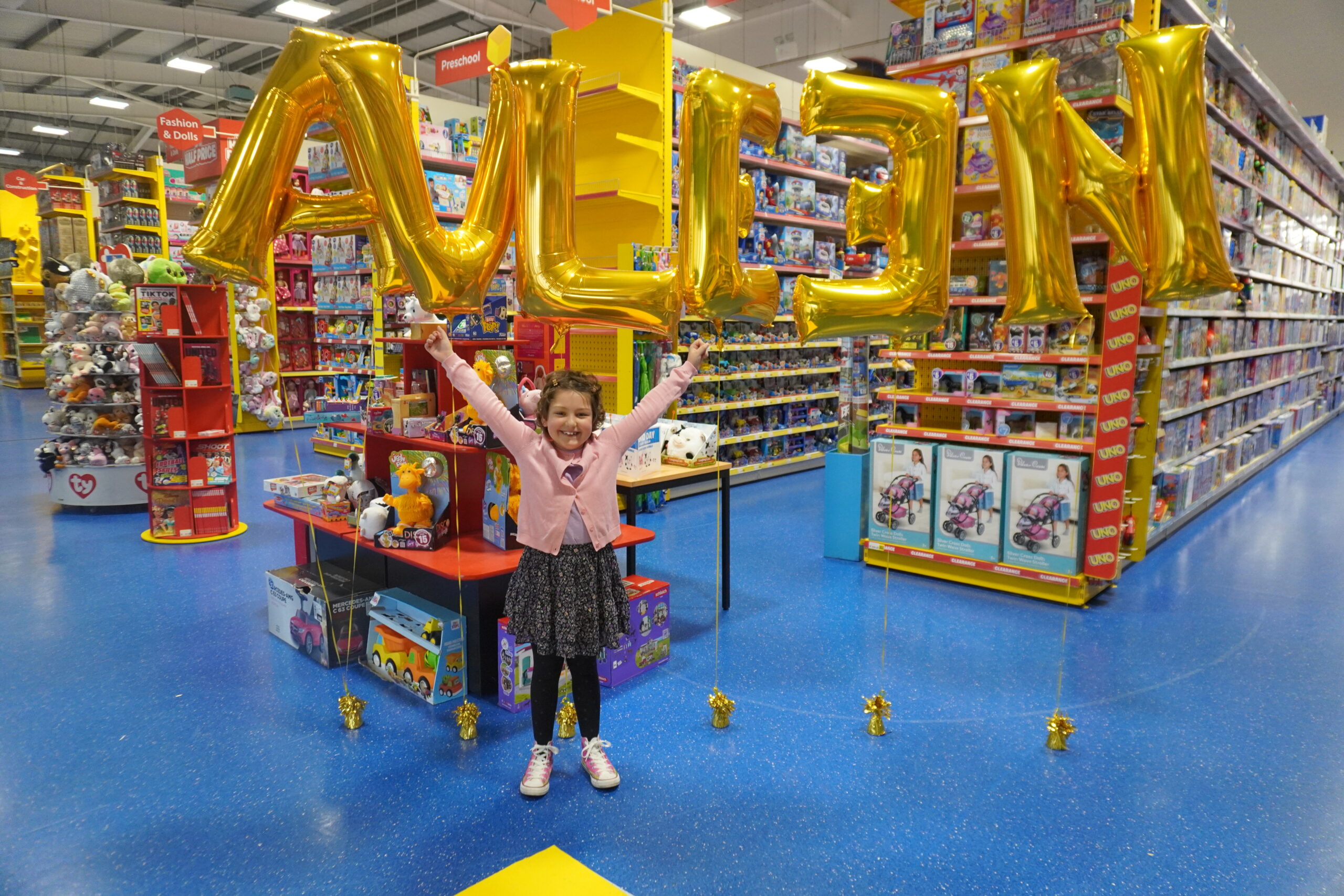 Little girl in a toy shop. There are balloons behind her which spell out AVLEEN. She is wearing a pink cardigan and is holding her arms up