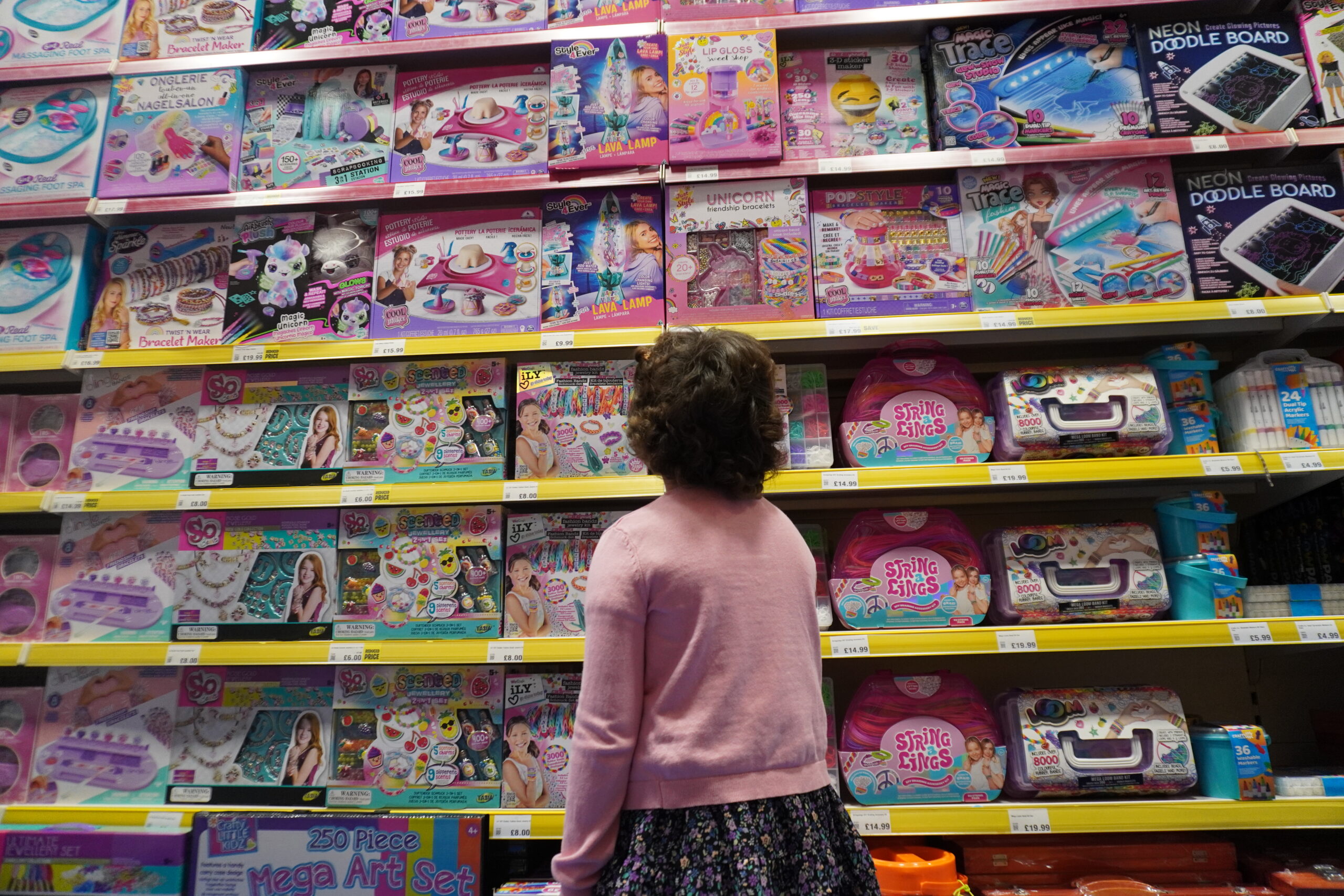 Picture of little girl with her back facing the camera. She is gazing up at all the different toys on the shelf of a toy shop.