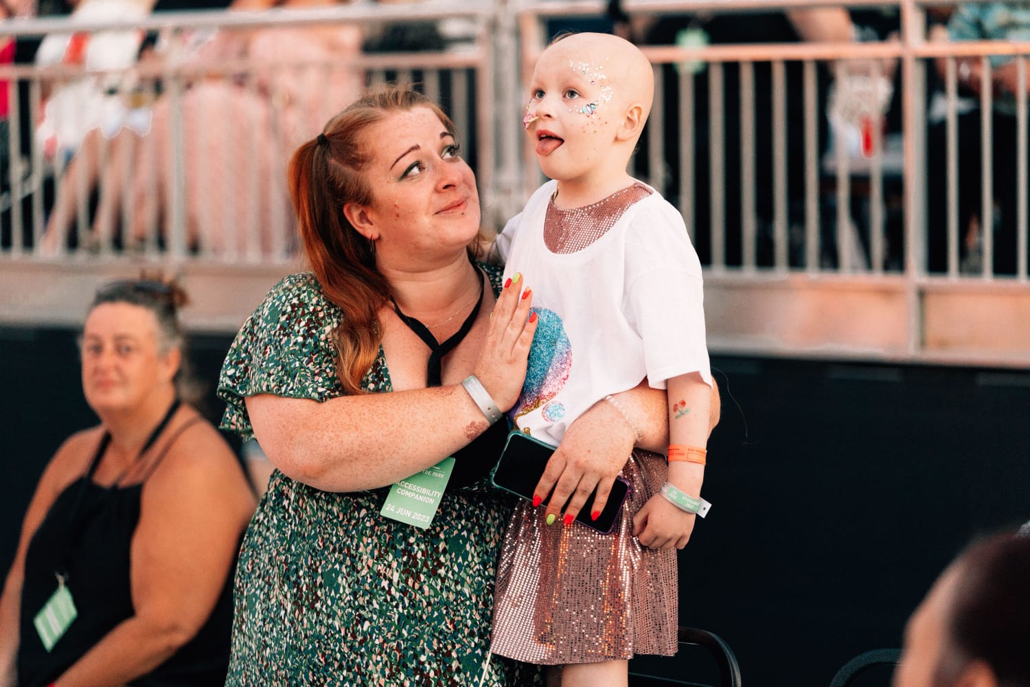 Little girl and her mum in the audience of a concert. The little girl is wearing a white t-shirt over a sparkly dress, and has no hair. The mum is wearing a green floral dress and gazing lovingly at her daughter.