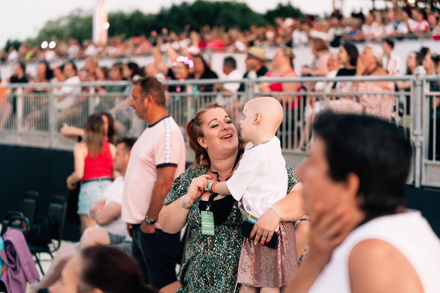 Little girl and her mum in the audience of a concert. The little girl is wearing a white t-shirt over a sparkly dress, and has no hair. The mum is wearing a green floral dress and smiling at her daughter.
