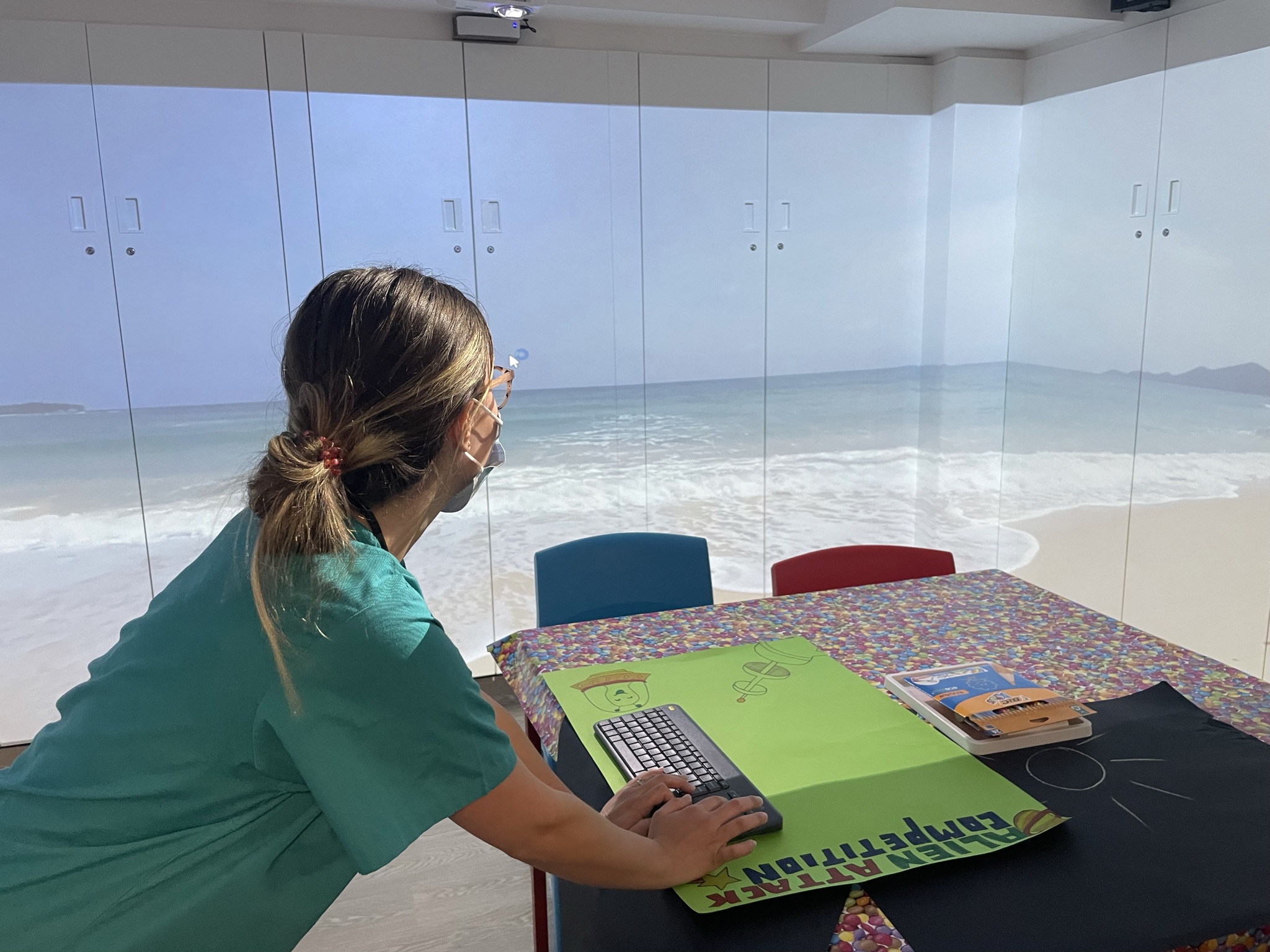 A nurse with her back to camera using immersive techology in a hospital room to show a sunny beach and sea on the walls