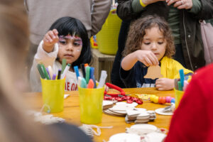 Two little girls sit at a craft table. They are both creating little pieces of art.