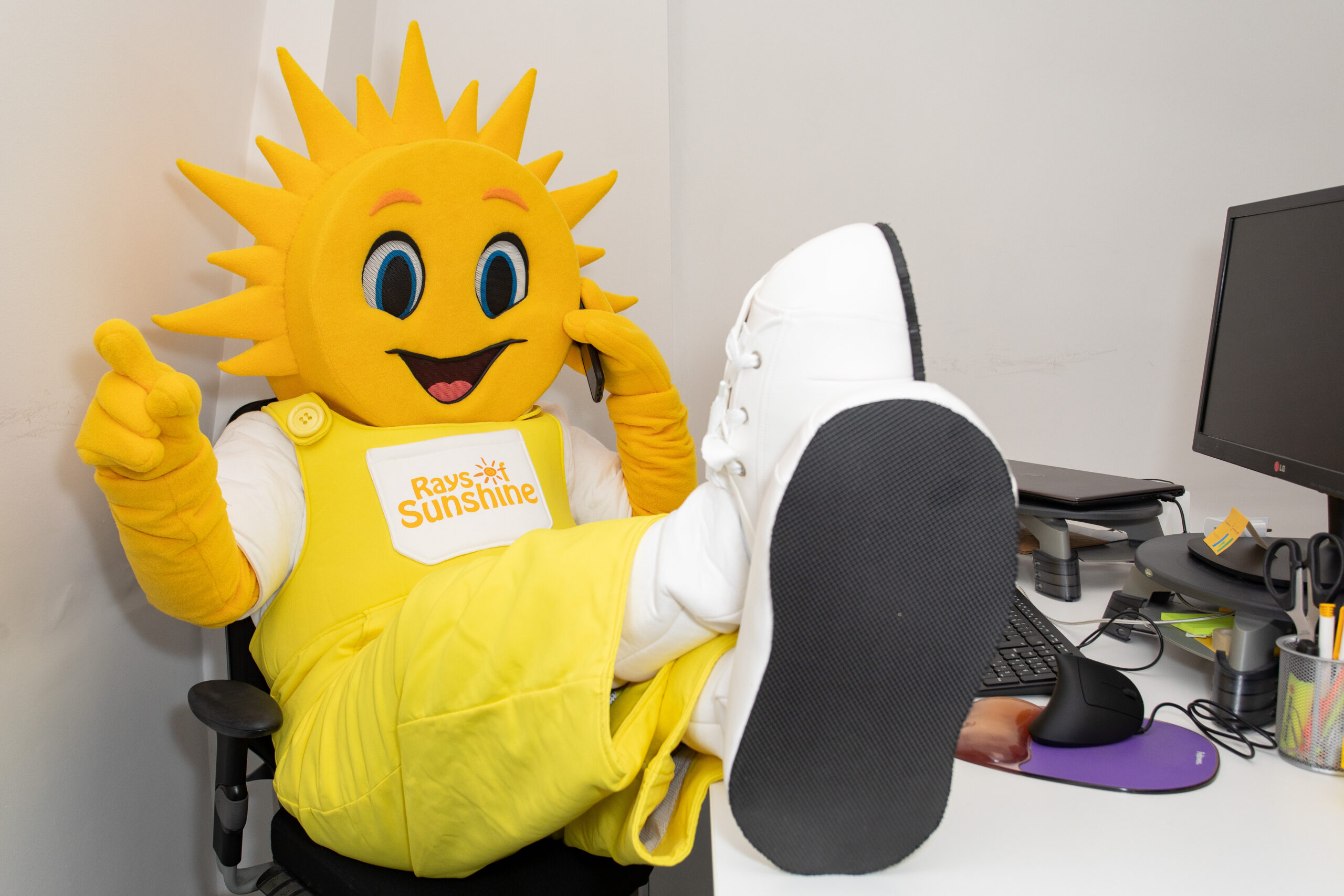 Rays of Sunshine mascot Sunny sat a desk pretending to be on the phone with their feet up at a desk