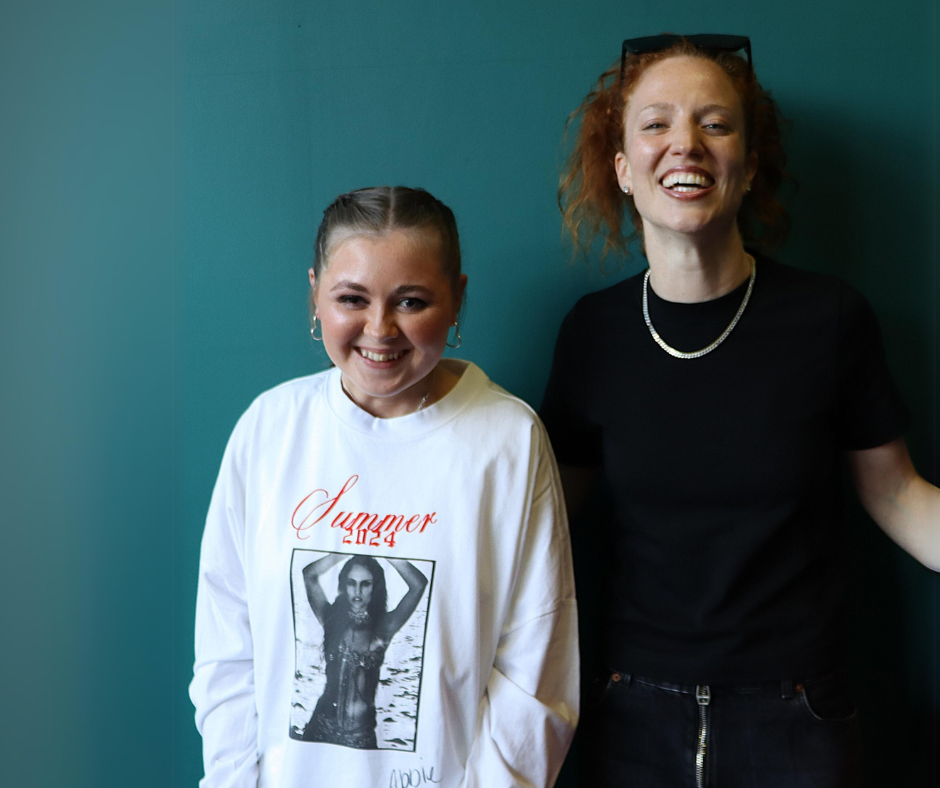 Teen girl smiles for the camera while stood next to woman who also smiles. The girl has french plaited hair, and hoop earrings, and wears a white long-sleeve top with a picture of a woman on the front. The woman is dressed all in black, with a silver chain. They are stood against a teal coloured wall