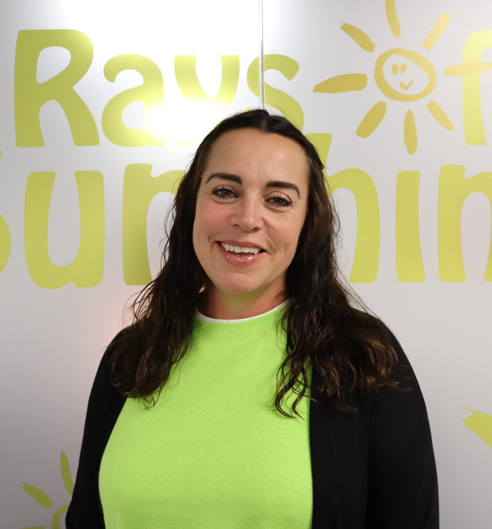 Headshot photo of a woman with dark curly hair. She is smiling. She wears a bright green top and black cardigan. She is stood in front of a window, which is decorated to say Rays of Sunshine in yellow writing.