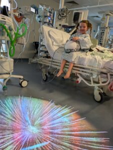 Little girl sits on hospital bed. She is wearing a hospital gown and pigtails, and cuddling a teddie. On the floor in front of her is a projector with lots of bright colours