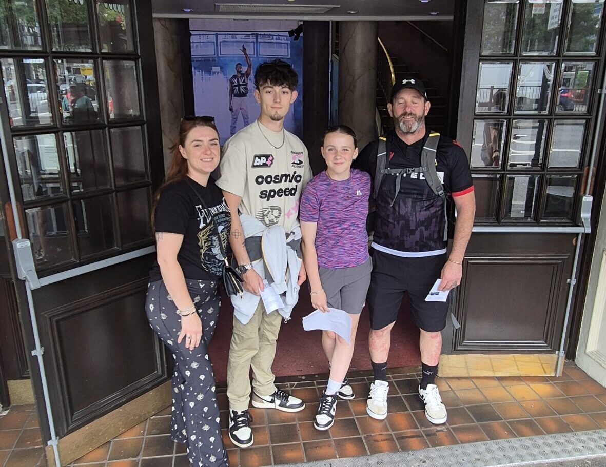 Family photo of four people - mum, dad, daughter and son. The son is in his late teens and is the tallest. They are stood in front of a wooden shopfront.