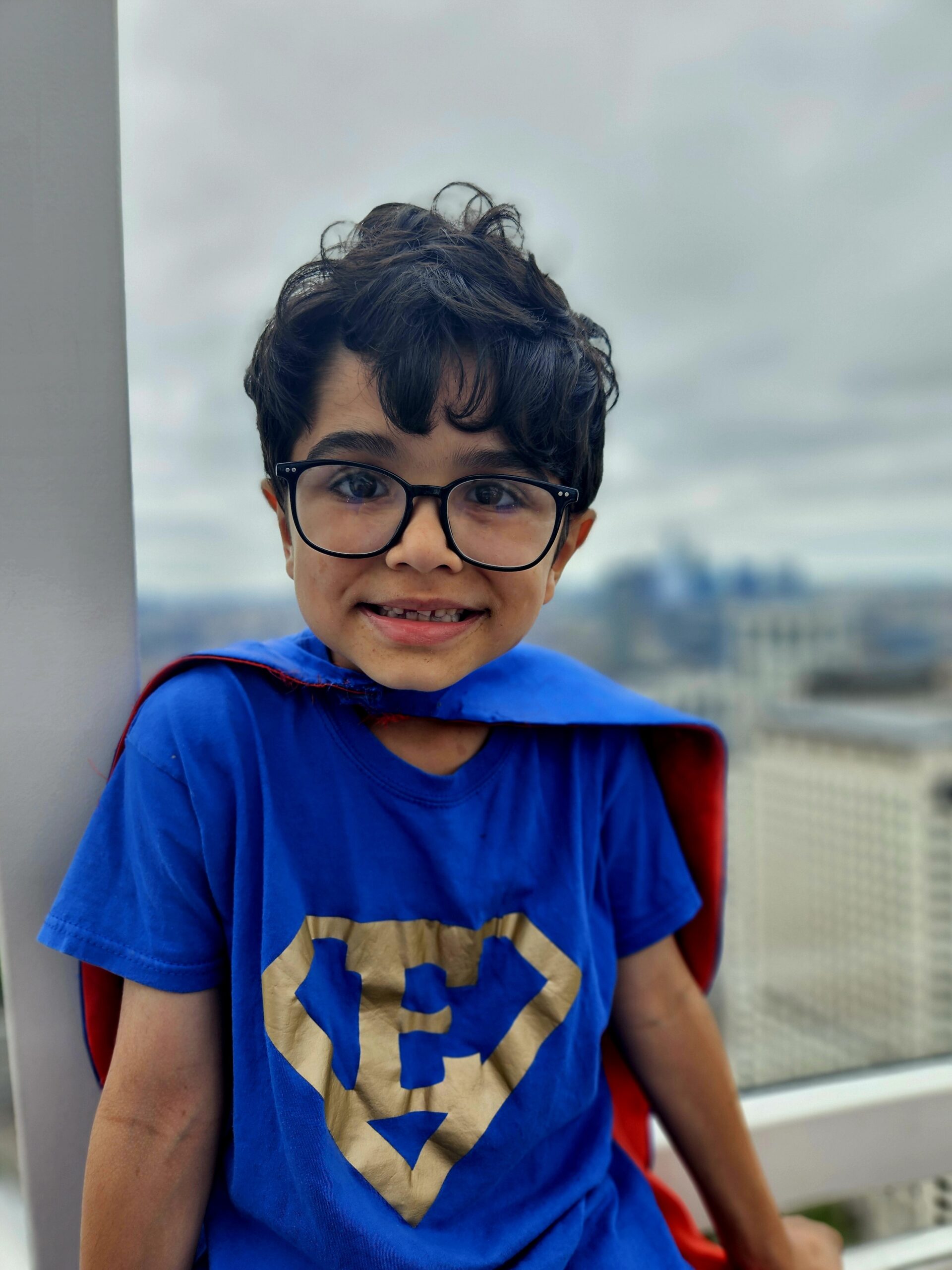 Little boy smiles for the camera. He has thick glasses and brown hair. He is wearing a blue t-shirt with a superhero emblem with an E on it.