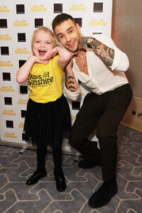 Little girl with yellow Rays of Sunshine t-shirt and black skirt and tights smiles for the camera while stood next to you man. He is wearing a white shirt and black trousers. They are both doing the same pose, with their hands under their chin. 