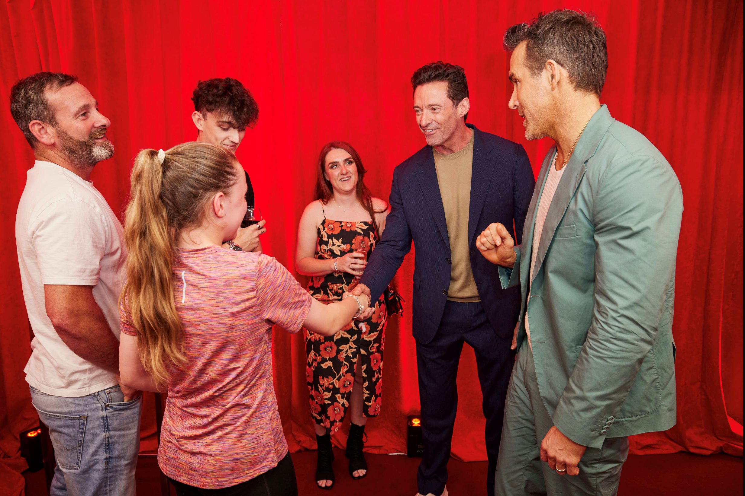 Six people in a group picture in front of a red curtain. None are look at the camera, but appear to be meeting for the first time. One of the men (Hugh Jackman) is shaking hands with a young girl.