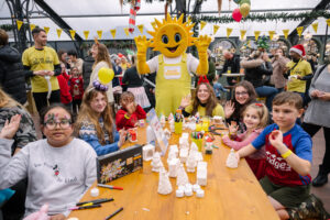 Group photo of several children sitting around a craft table at a Christmas party. Everyone is very smily, and in the background is a sunshine-head mascot 