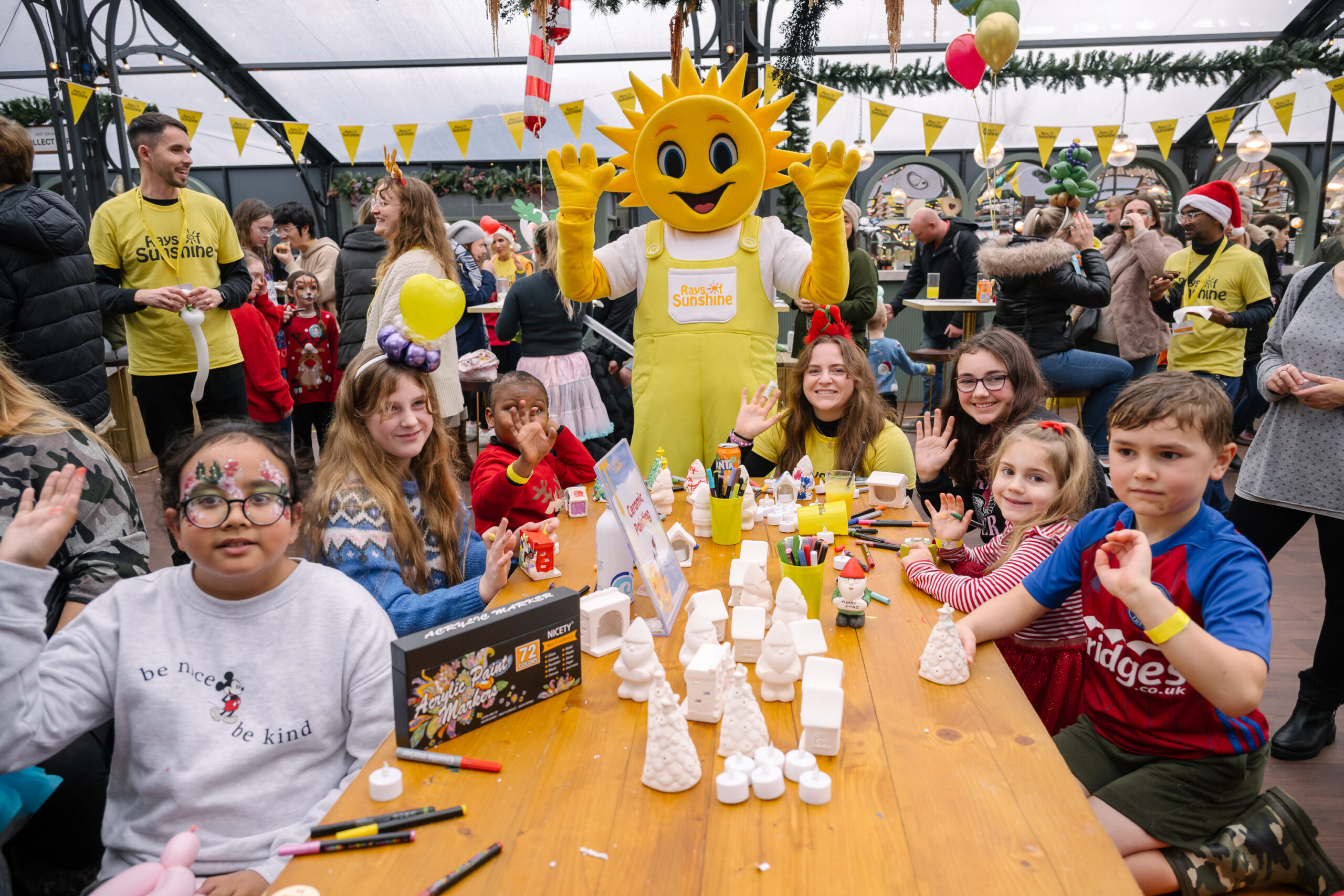 Group photo of several children sitting around a craft table at a Christmas party. Everyone is very smily, and in the background is a sunshine-head mascot