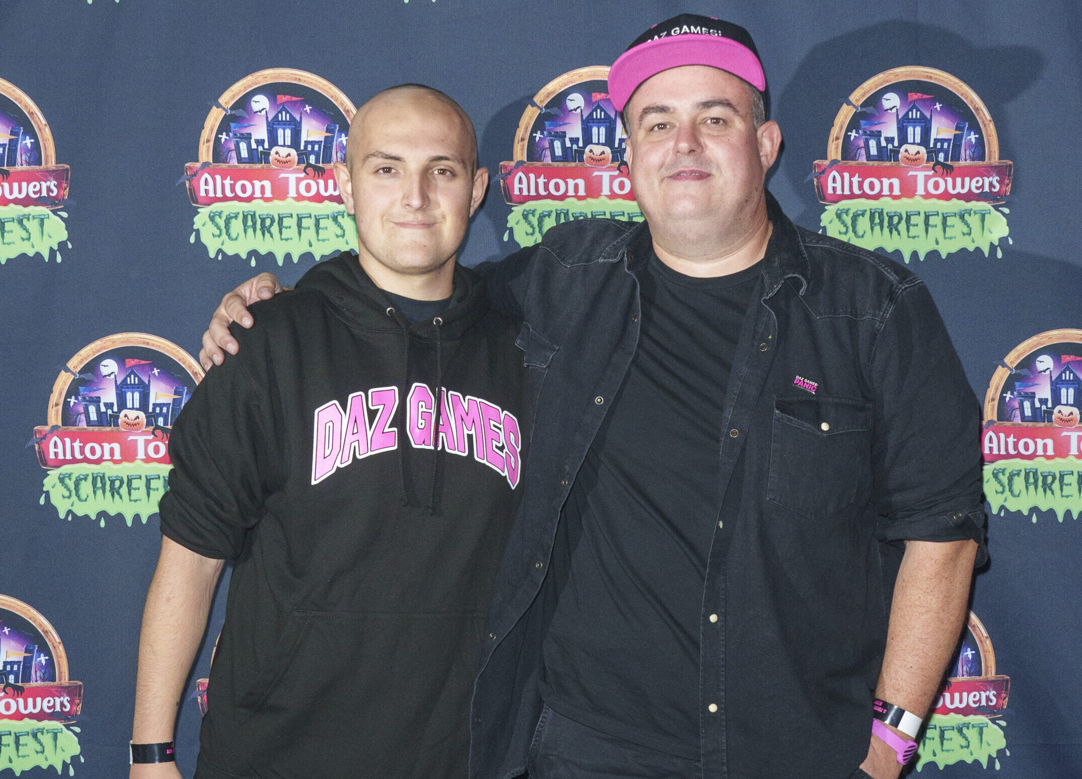 Two men standing in front of a step and repeat board featuring the Alton Towers logo. The younger of the two men is wearing a black hoodie which reads Daz Games, while the older is wearing an open black shirt and t-shirt and a baseball cap with a pink brim.