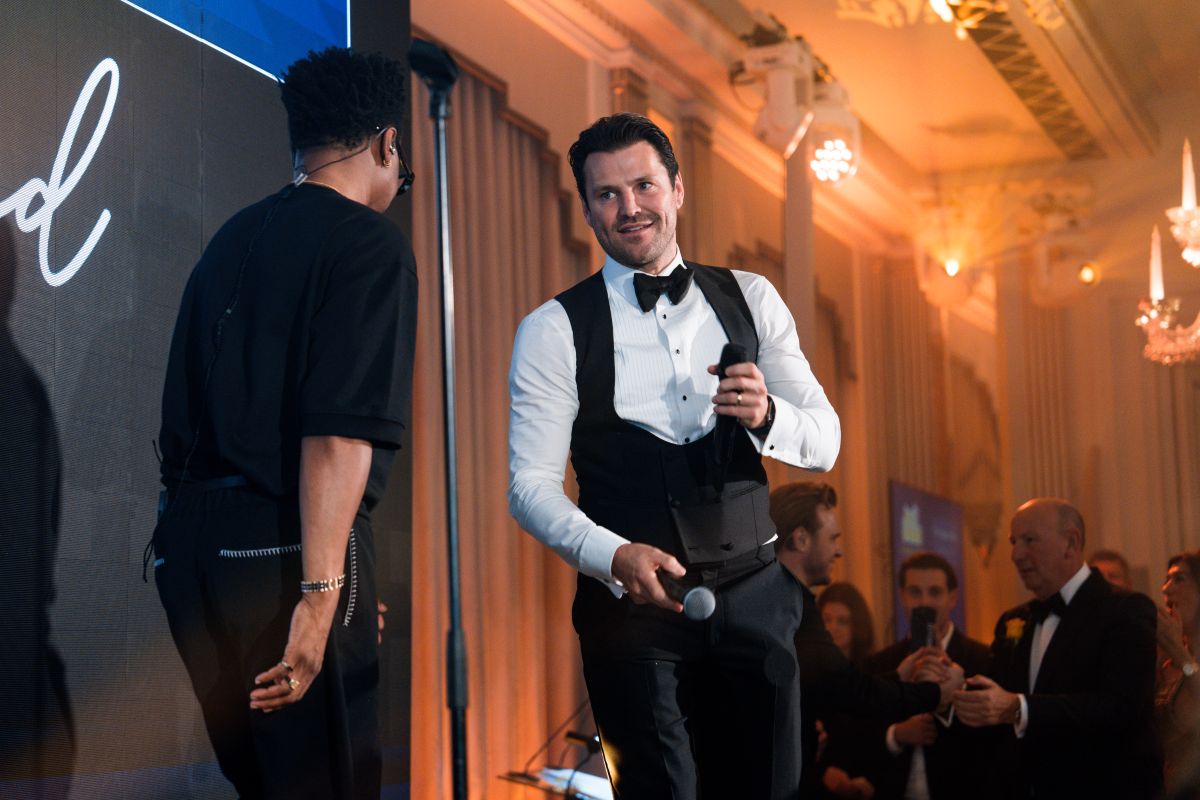 Man in black waistcoat and white shirt smiles while on stage