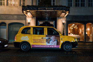 Bright yellow taxi parked in front of fancy hotel. The driver's door of the taxi has a little girl's face on it