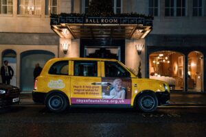 Bright yellow taxi parked in front of fancy hotel. The driver's door of the taxi has a little girl's face on it