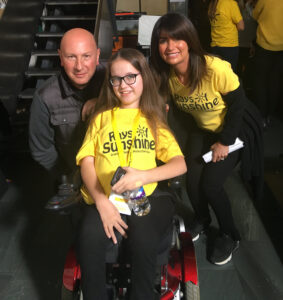 A young girl in a wheelchair wearing a yellow Rays of Sunshine T-shirt smiles with two adults, one in a grey vest and the other in a matching yellow T-shirt, at an indoor event