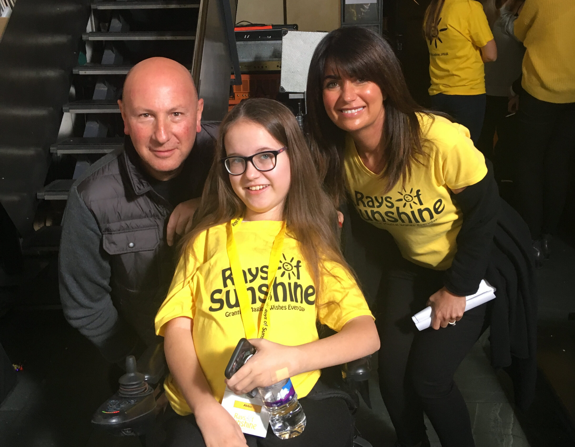 A young girl in a wheelchair wearing a yellow Rays of Sunshine T-shirt smiles with two adults, one in a grey vest and the other in a matching yellow T-shirt, at an indoor event