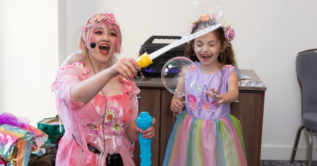 A woman dressed as a princess is waving a bubble wand next a girl in a colourful dress smiling