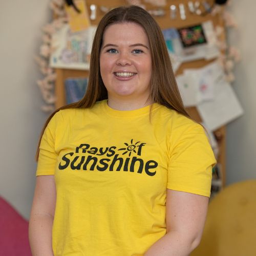 Rhea is wearing a Rays of Sunshine top. She is standing. Rhea has brown hair