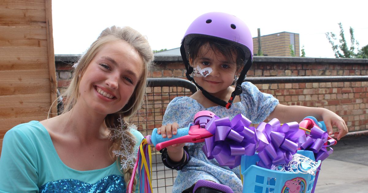 A princess is smiling next to a girl wearing a helmet sat on a new bike