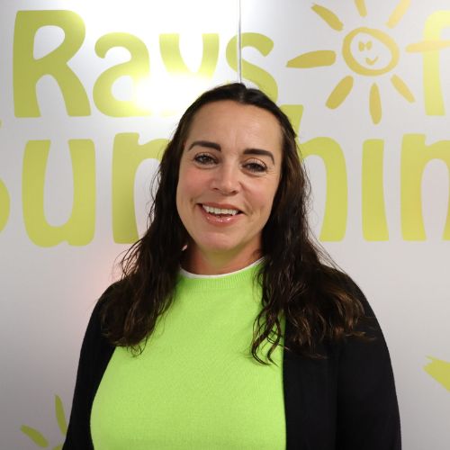 Headshot photo of a woman with dark curly hair. She is smiling. She wears a bright green top and black cardigan. She is stood in front of a window, which is decorated to say Rays of Sunshine in yellow writing