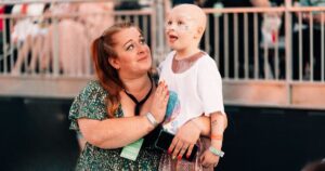 A mother holding up and looking up at their child. The child has no hair and is wearing sparkly clothes