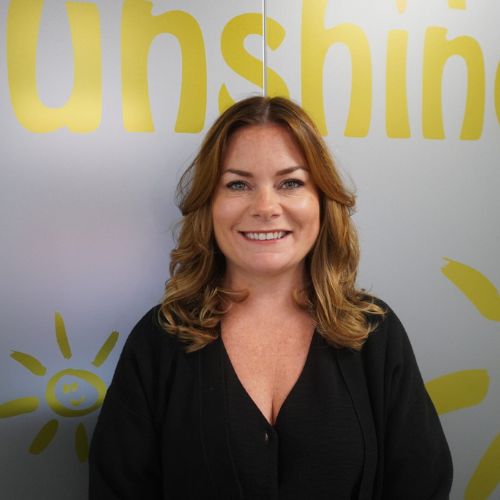 Amy is smiling at the camera she has long ginger hair and is standing in front of a Rays of Sunshine logo