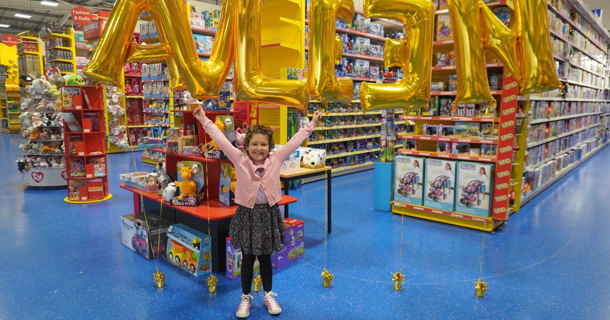A young girl standing in a toy store smiling with her arms in the air in front of balloons