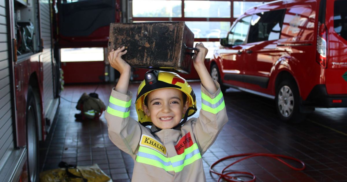 A young boy wearing a firefighter uniform and holding a box on his head