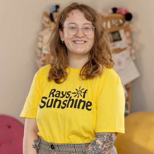 Molly is wearing a yellow Rays of Sunshine t-shirt. She is standing. Molly has brown hair.