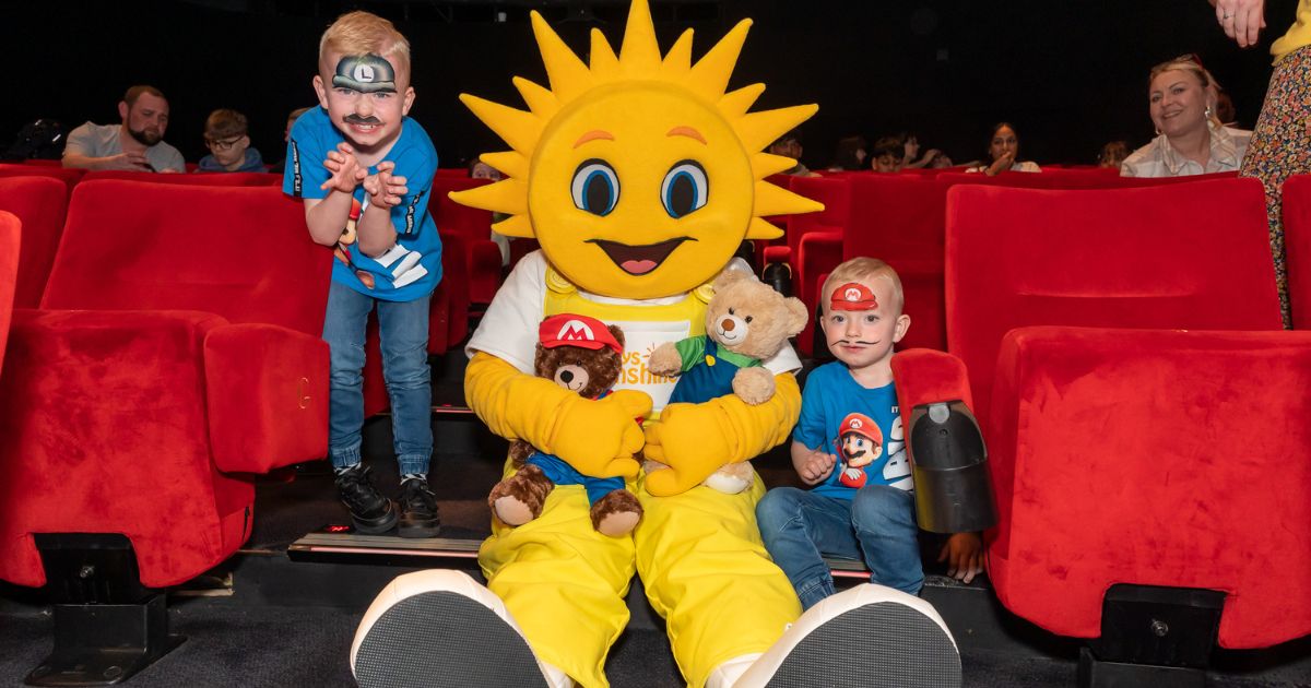 Rays of Sunshine mascot sat down in a movie theatre with two children with face paint on holding teddies