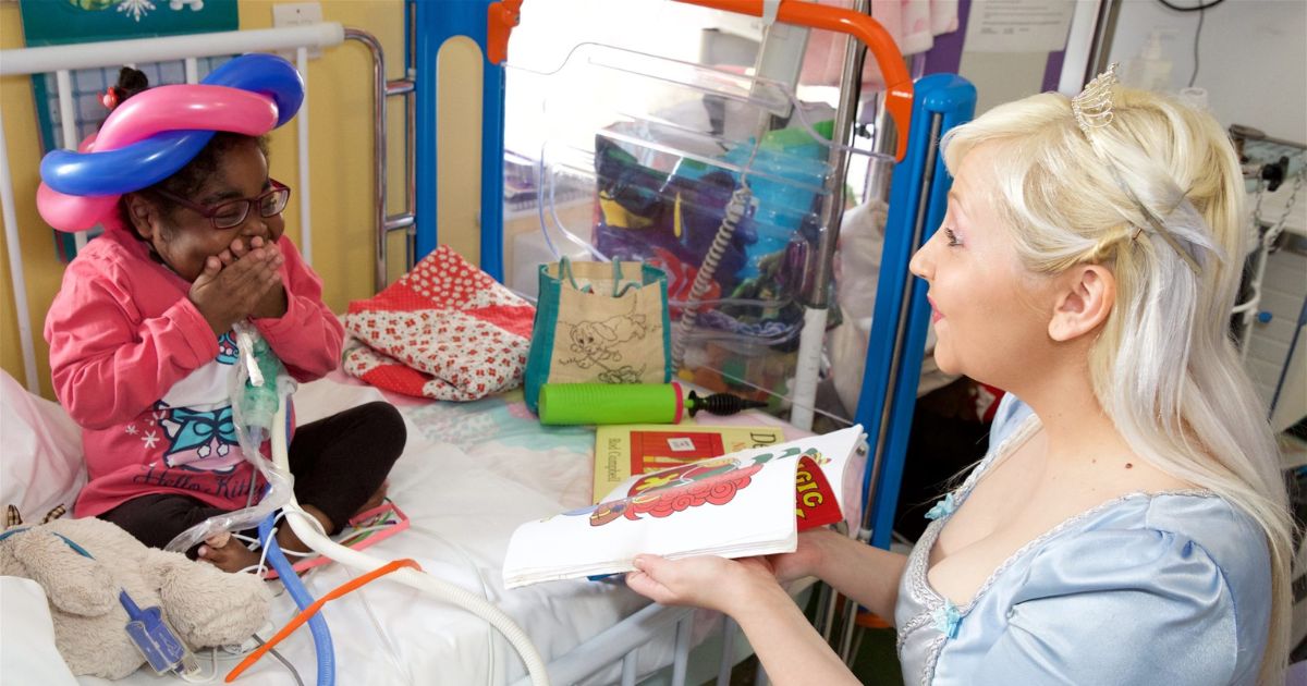 A woman dressed a princess reading a book to a child in a hospital bed who is laughing and covering her mouth