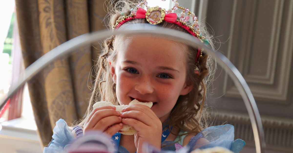 A child smiling to camera wearing a princess headband eating a sandwich