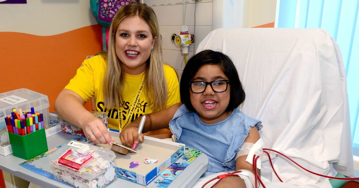 A woman wearing a Rays of Sunshine yellow t-shirt sat next to a child in a hospital bed drawing. Both are smiling.