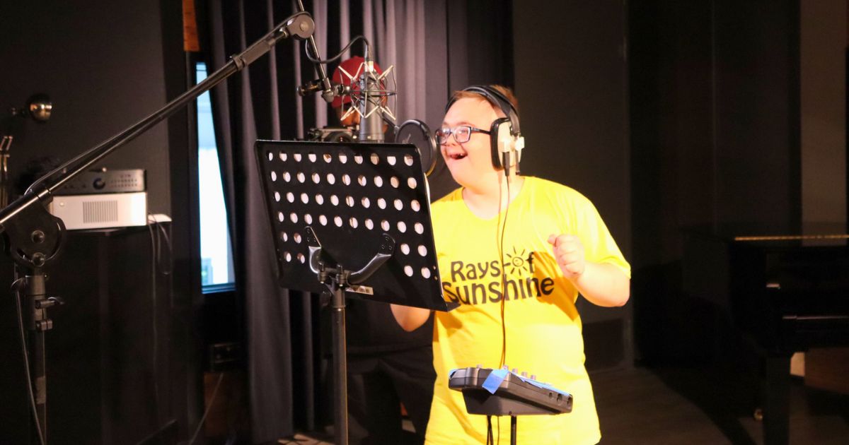 A young person with downs syndrome wearing a yellow Rays of Sunshine t-shirt singing into a microphone