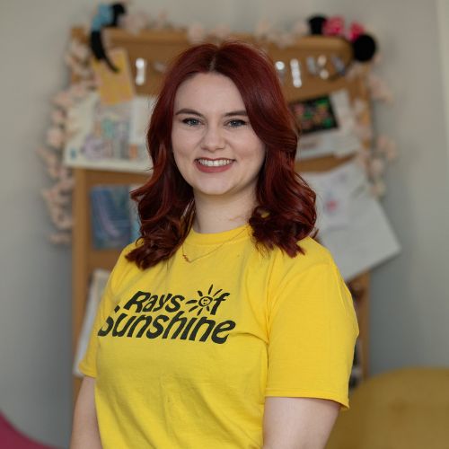 Kathryn is wearing a yellow Rays of Sunshine t-shirt. Kathryn is standing and has red hair